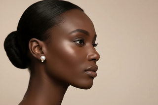 a woman with dark skin and styled hair against a beige background wearing a pearl and diamond stud earrings