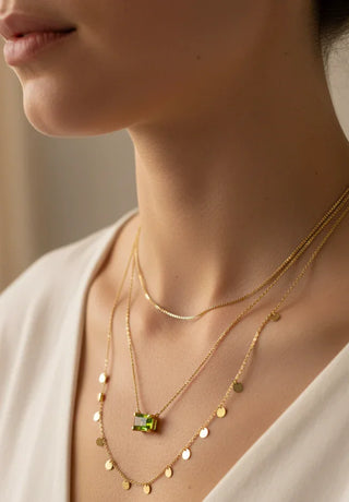 Close-up of a person wearing gold necklaces with a green gemstone on a neutral background