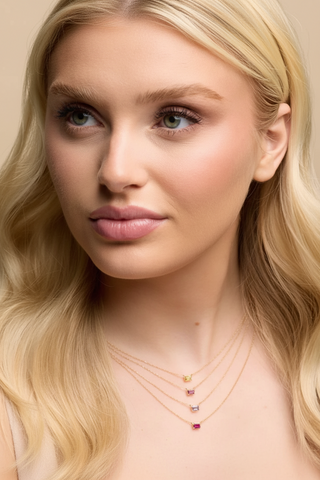 A close-up portrait of a blonde woman wearing delicate layered necklaces with small colorful gemstones on a soft beige background.