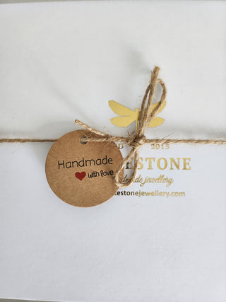 A close-up of a white jewellery box or card with gold text reading 'WHITESTONE handmade jewellery' and 'whitestonejewellery.com', tied with natural twine and a round kraft paper tag that says 'Handmade with love' with a small red heart.