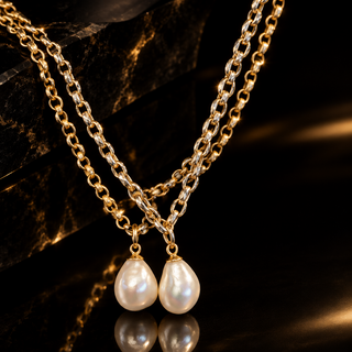A double-strand gold chain necklace with teardrop-shaped white freshwater pearls, displayed on a dark reflective surface with a polished black crystal prop.