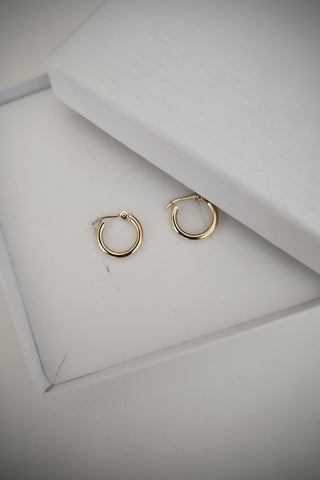 A pair of small gold hoop earrings displayed on a white jewelry box lid, with part of a white branded box visible in the background.