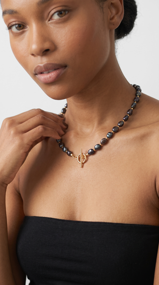 A close-up of a model wearing a dark grey beaded necklace with a gold-toned toggle clasp, paired with a black strapless top against a light grey background.