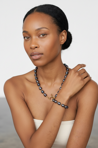 A woman wearing a dark gray beaded necklace with a gold toggle clasp, styled with a white strapless top against a light gray background.