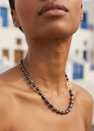 A close-up lifestyle shot of a model wearing a dark grey and black beaded necklace with a gold-tone toggle clasp, set against a blurred background of white buildings with blue accents.