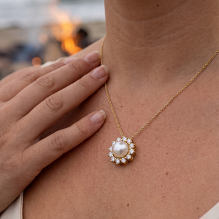 A close-up lifestyle shot of a woman wearing a delicate gold chain necklace with a central white pearl surrounded by small clear stones, with her hand resting on her chest against a softly blurred background.