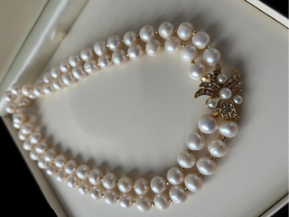 A double-strand pearl necklace with a gold butterfly-shaped charm embellished with small clear stones, displayed inside a cream-colored jewelry box.