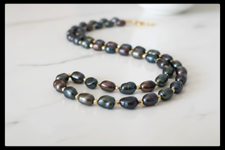 A baroque pearl necklace with iridescent dark green, purple, and blue-grey beads separated by small gold spacers, displayed on a white marble surface.