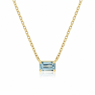 A delicate yellow gold chain necklace featuring a rectangular emerald-cut light blue aquamarine gemstone pendant in a prong setting, shown against a clean white background with a soft reflection below.