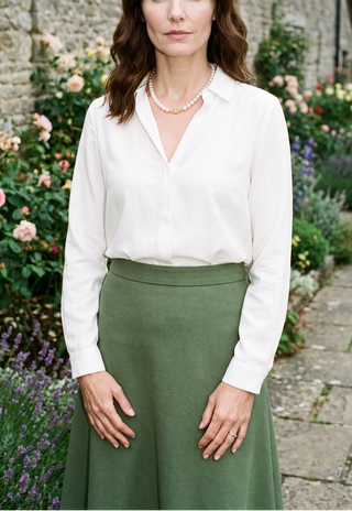 A woman wearing a white long-sleeve button-up blouse and an olive green skirt, standing outdoors in a garden setting with flowers and a stone wall behind her.