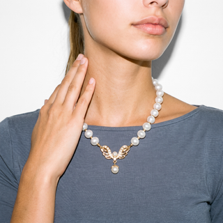A close-up of a model wearing a pearl necklace with a gold leaf pendant, styled with a slate blue top against a clean white background.