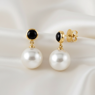 A pair of gold drop earrings with round black onyx tops and large white freshwater pearls hanging below, displayed on a draped white satin fabric background.