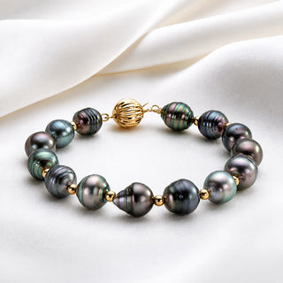 A beaded pearl bracelet featuring dark iridescent baroque pearls with gold spacer beads and a textured gold clasp, displayed on a draped white satin fabric.
